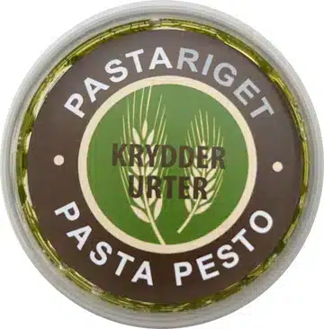 Pastariget, Krydderurter Pesto Rundt låg med grøn/brun design, urter illustrationer. Tekst: "PASTARIGET KRYDDERURTER PASTA PESTO".